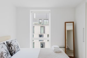 A hotel room with a bed, a mirror, and a view of a building with multiple floors.