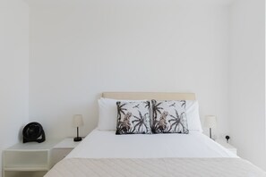 A neatly made bed with a white headboard, two white nightstands with lamps, and a single decorative pillow with a palm tree design.