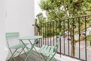 Ein Balkon mit Tisch und zwei Stühlen, der auf eine Straße mit parkenden Autos und Bäumen blickt.