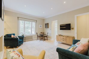 Ein geräumiges Wohnzimmer mit einer Couch, einem Esstisch mit Stühlen, einem an der Wand montierten Fernseher und einem großen Fenster, das für natürliches Licht sorgt.