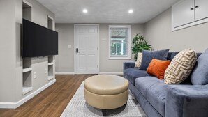 A living room with a flat-screen TV mounted on a white wall, a grey sofa with patterned cushions, a round ottoman, and a window with blinds. - Charming 1BR apt -close to public transit -has parking -30min drive to BOS/Salem (Reading)