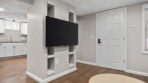 A modern living room with a flat-screen TV mounted on a white wall, a wooden floor, and a white door. - Charming 1BR apt -close to public transit -has parking -30min drive to BOS/Salem (Reading)