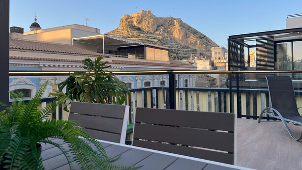 Un balcón con sillas y una mesa de madera, con vistas a una montaña y edificios.