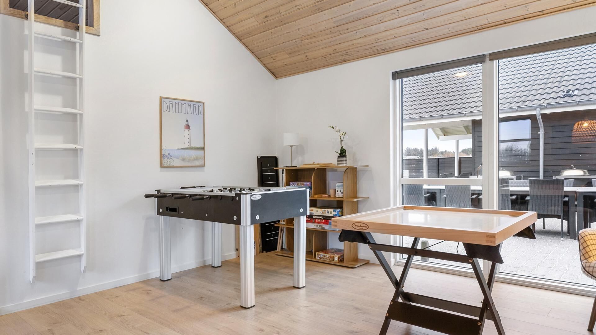 A room with a foosball table, a bookshelf, a painting of a lighthouse, and a view of the outside.