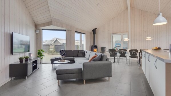 A modern living room with a grey sectional sofa, a flat-screen TV mounted on the wall, and a large window offering a view of the outside.