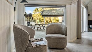 Un interior modern amb dues cadires tapissades, una taula amb una revista, i vista a una zona exterior amb taula de picnic i arbres.