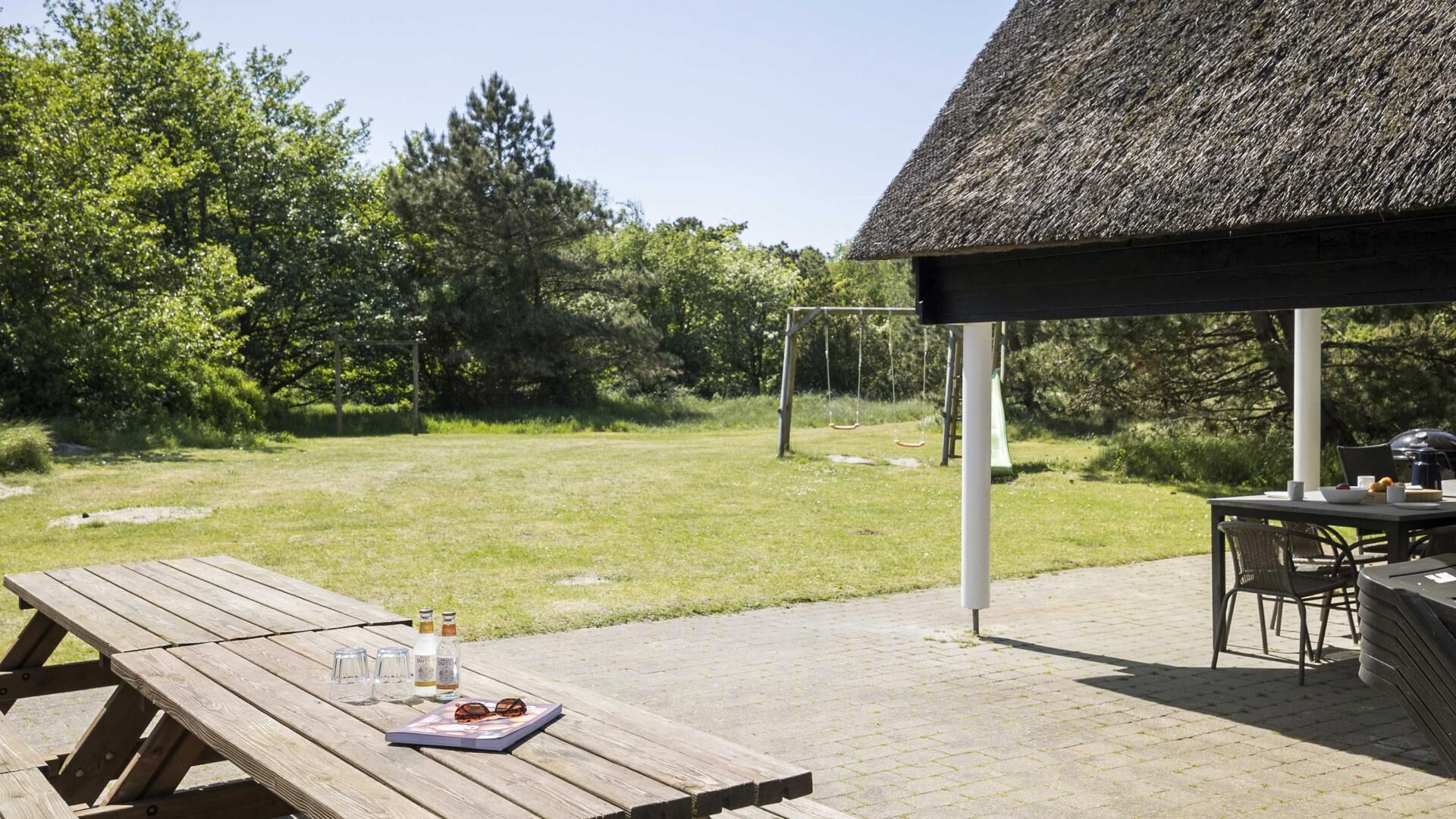 Een houten picknicktafel met een fles, glazen en zonnebril, een overdekte zitplek en een gazon met bomen.