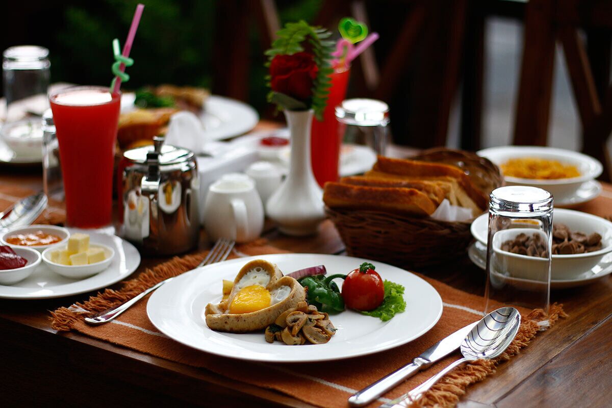 Una escena de desayuno con un plato de huevos, champiñones y verduras de hoja, un vaso de bebida roja, y varias salsas y pan sobre la mesa.
