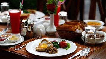 Una escena de desayuno con un plato de huevos, champiñones y verduras de hoja, un vaso de bebida roja, y varias salsas y pan sobre la mesa.