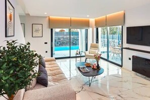 Ein modernes Wohnzimmer mit einer Couch, einem Couchtisch und einem Fernseher. Ein großes Fenster bietet Blick auf einen Pool und die Skyline der Stadt.