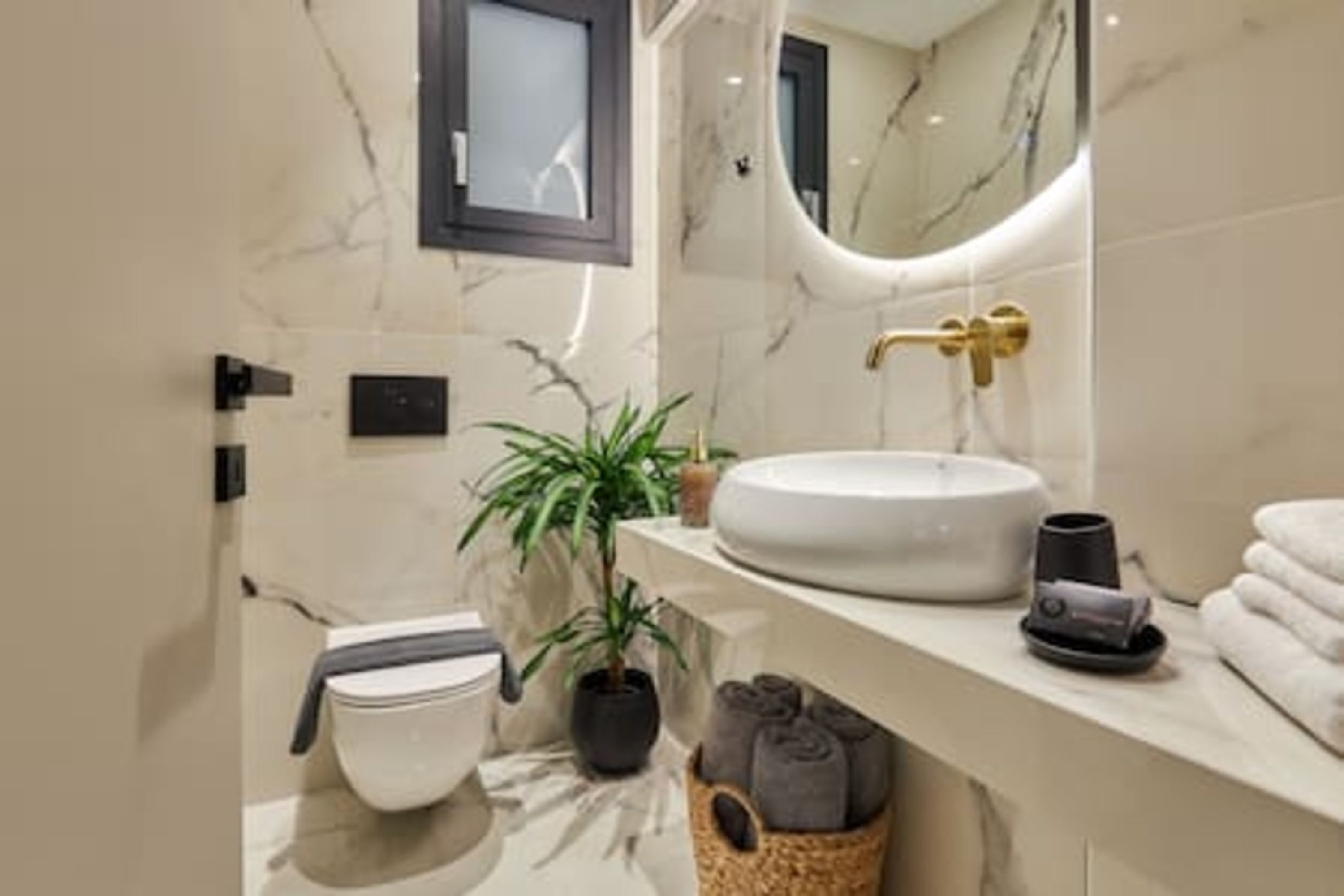A modern bathroom with a marble sink, a gold faucet, a potted plant, and neatly rolled towels.