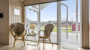 Une pièce comprenant deux fauteuils en osier, une petite table et une lampe. Les murs de la pièce sont en bois et une grande porte-fenêtre coulissante donne sur un espace extérieur.