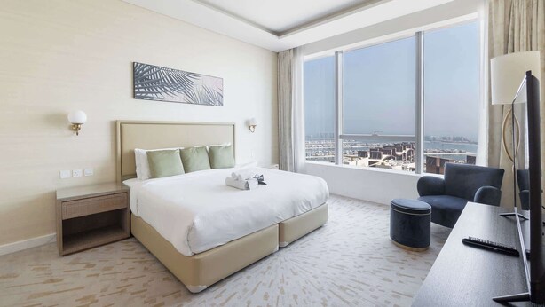 Ein Hotelzimmer mit einem großen Bett, einem Schreibtisch mit Stuhl, einem Fernseher und Blick auf die Stadt durch große Fenster.