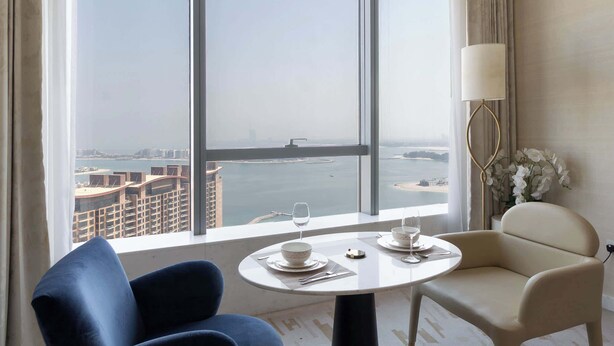 Habitación de hotel con un ventanal que ofrece vistas a un paisaje urbano y una masa de agua. Hay una mesa de comedor redonda con dos sillas, una lámpara y un jarrón con flores.
