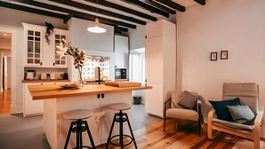Ein moderner Wohnraum mit Küchenbereich, Holzboden, einer Bar mit Hockern und einer Sitzecke mit Sessel und Kissen.