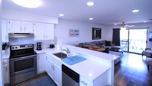 A modern kitchen with white cabinetry, a built-in oven, and a dishwasher. There is a dining area with a table and chairs, and a living room with a sofa and a TV. - Bayview Freedom (Orange Beach)