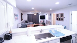 A modern kitchen with a central island, a dining area with a table and chairs, and a living room with a sofa and a TV. - Bayview Freedom (Orange Beach)