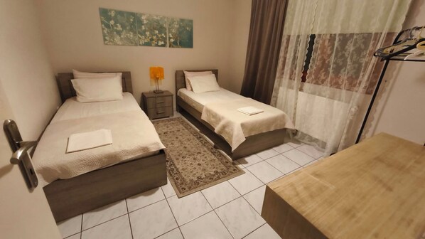 A hotel room with two single beds, a small table, a nightstand, a lamp, and a painting on the wall.