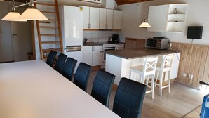 A compact kitchen with white cabinets, a microwave, and a stove. There is a dining area with a long table and chairs. - VillaVilla 127 - Öland, Sverige (Köpingsvik)
