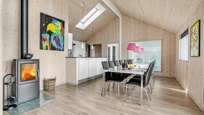 A modern cabin interior with a wood-burning stove, a dining area with a table and chairs, a kitchen with white cabinets, and artwork on the walls. - VillaVilla 299 - Marielyst, Lolland/Falster/møn (Idestrup)