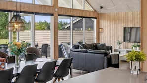 A modern living room with a large sofa, dining table, and chairs. There is a TV mounted on the wall and a large window providing natural light.