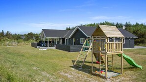 Een speeltuin met een glijbaan en klimconstructie, een overdekt zitgedeelte en een gebouw met een donker dak en ramen.