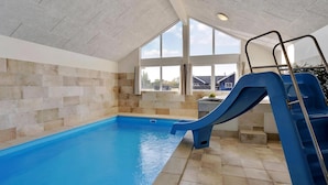 Une piscine intérieure avec un toboggan bleu, des murs en pierre et de grandes fenêtres.