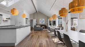 Une cuisine moderne avec un îlot central, un coin repas avec une table et des chaises blanches, et un salon avec un canapé et un tableau au mur.