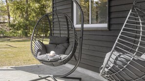 A hanging chair with cushions and a ball of yarn, positioned on a paved patio next to a building with a window.