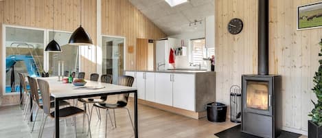 A modern kitchen with wooden walls, a dining area with a table and chairs, a visible clock, and a wood-burning stove.