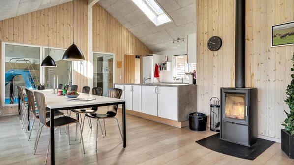Een moderne keuken met houten wanden, een eethoek met een tafel en stoelen, een zichtbare klok en een houtkachel.