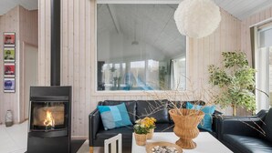 A cozy living room with a fireplace, a black leather sofa, a white coffee table with decorative items, and a large window offering a view of the outdoors.