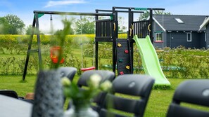 A playground with a slide and climbing area, surrounded by a grassy field and a house in the background.