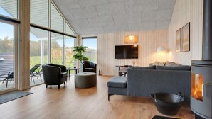 Een moderne woonkamer met een open haard, een aan de muur bevestigde televisie en een groot raam met uitzicht op een tuin.