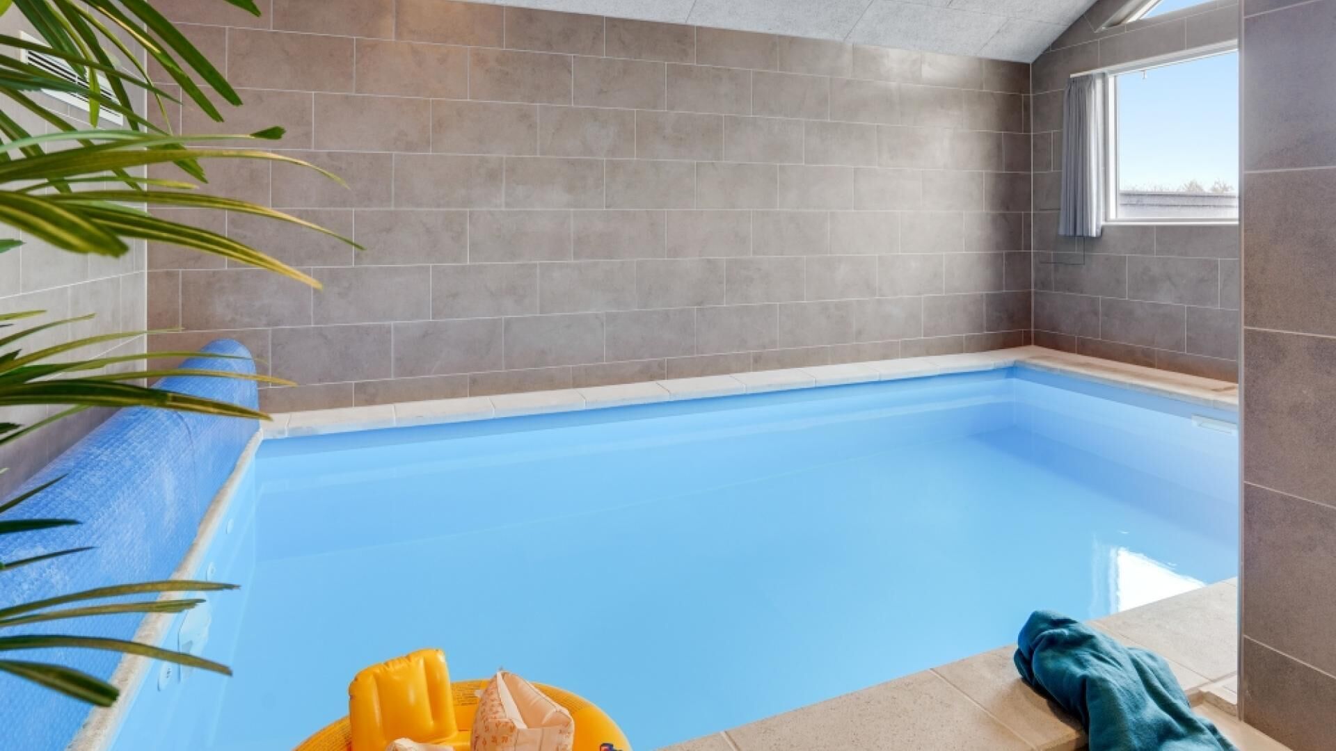 Una piscina interna con superficie d'acqua blu, circondata da pareti piastrellate di colore grigio e una finestra su un lato.