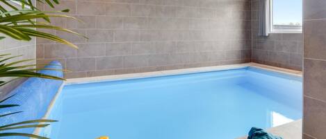 Una piscina interna con superficie d'acqua blu, circondata da pareti piastrellate di colore grigio e una finestra su un lato.