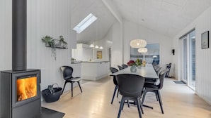 A modern interior with a wood-burning stove, a dining area with chairs and a table, and a kitchen in the background.
