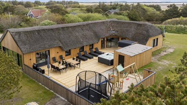 Een houten chalet met rieten dak, buitenzitplaatsen, een jacuzzi en een trampo.