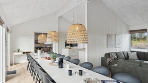 Una cocina moderna con muebles blancos, una zona de comedor con una mesa larga y sillas, y un sofá gris con cojines.
