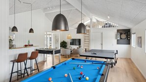Amplia sala con mesa de billar, mesa de fútbol de mesa y mesa de ping pong. Hay dos taburetes de bar y un televisor montado en la pared.