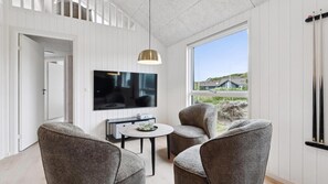 A modern living room with a TV, two grey armchairs, a small table with glasses, and a large window with a view of houses.