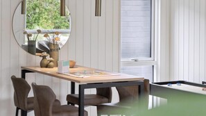 Une pièce avec une table de billard, des chaises, un miroir et une fenêtre avec des stores.