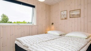 A room with wooden panel walls, a bed with white bedding and green pillows, two framed artworks, and a window with a view of greenery.