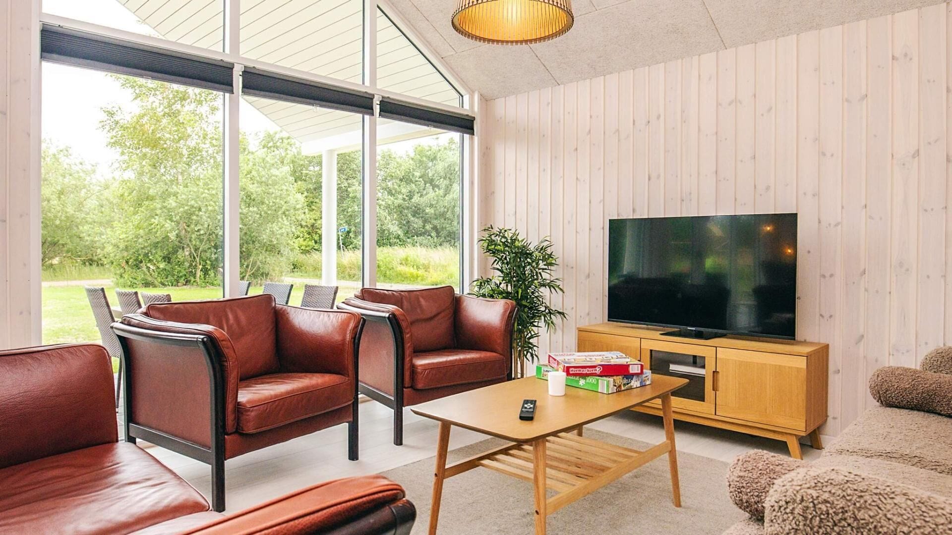 A modern living room with leather sofas, a wooden coffee table, a TV on a wooden cabinet, and large windows offering a view of greenery.