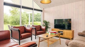 Een moderne woonkamer met leren banken, een houten salontafel, een televisie op een houten kast en grote ramen met uitzicht op groen.