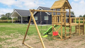 Una estructura de juego de madera con un tobogán y columpios, una zona de estar cubierta y una casa al fondo.