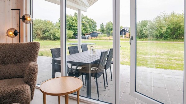 A modern living room with a sofa, a small round table, and a dining area with a table and chairs. Large glass doors lead to an outdoor patio with a lawn and a house in the background.