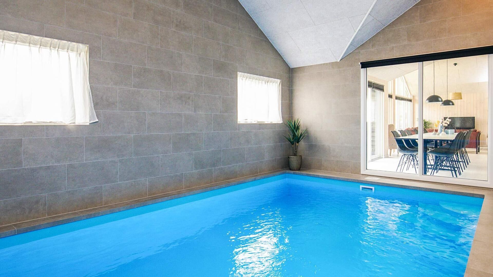 An indoor swimming pool with grey tiled walls, a potted plant, and a dining area visible through a glass door.