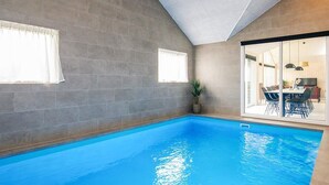Una piscina interior con paredes de baldosas grises, una planta en maceta y un área de comedor visible a través de un vidrio.