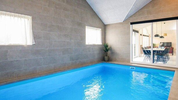 An indoor swimming pool with grey tiled walls, a potted plant, and a dining area visible through a glass door.